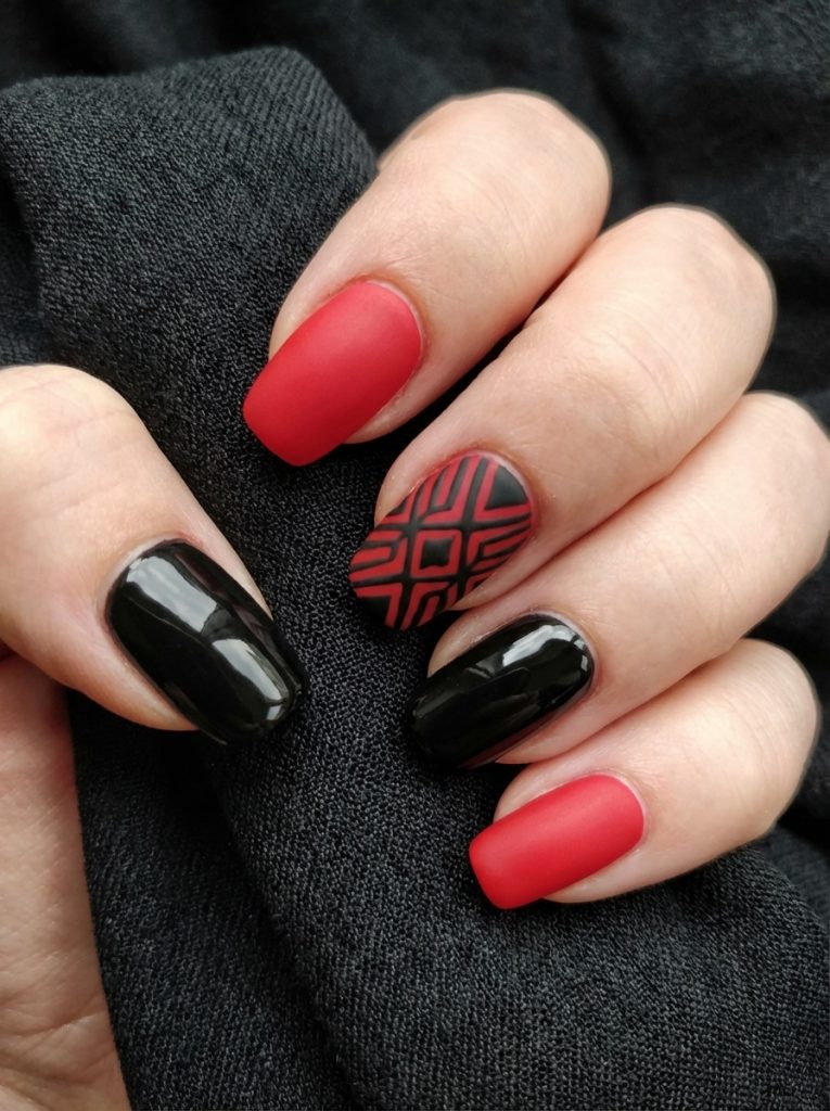 Hand gripping textured black fabric reveals red and black wedding nails in matte crimson and high-gloss black, featuring a sharply detailed geometric accent design.