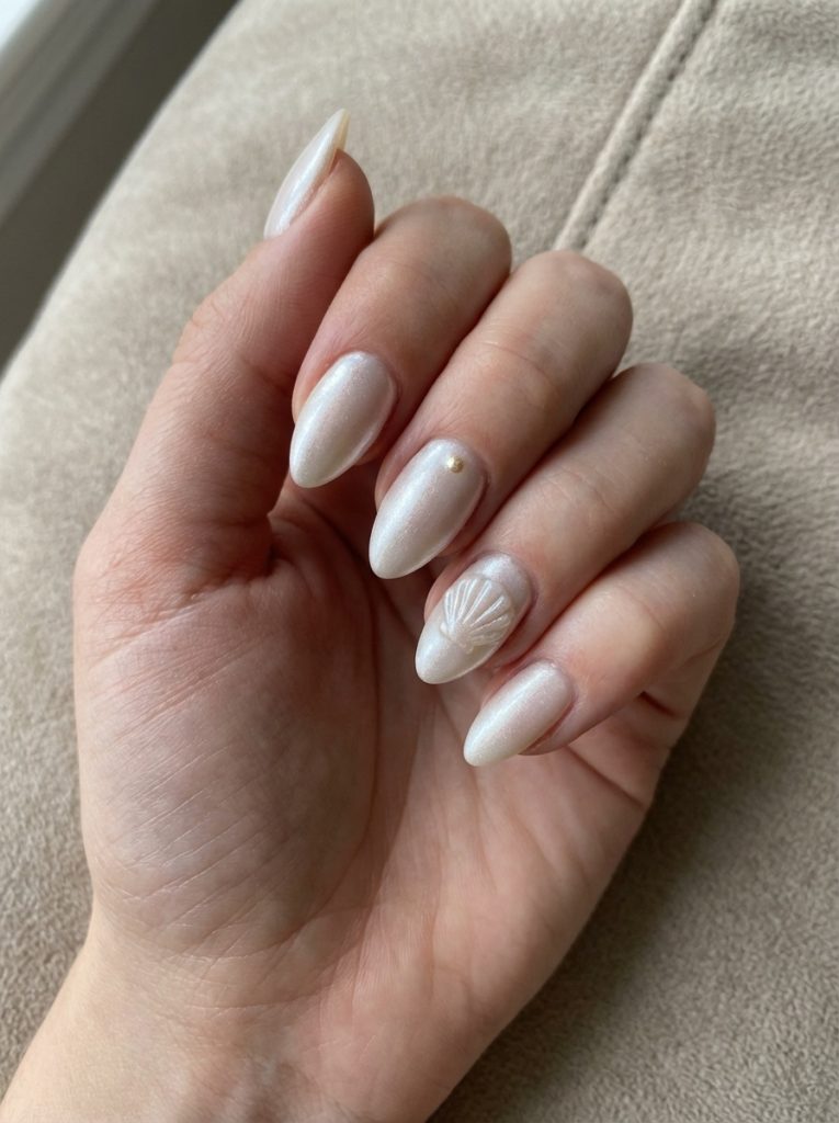 Almond-shaped pearl wedding nails painted champagne ivory with subtle shimmer, highlighted by a raised shell pattern and a delicate gold embellishment on one nail.