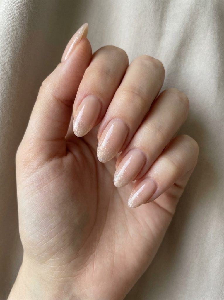 A relaxed hand pose reveals nude wedding nails in glossy blush nude, enhanced by fine pearly shimmer that becomes slightly stronger near the almond-shaped tips.