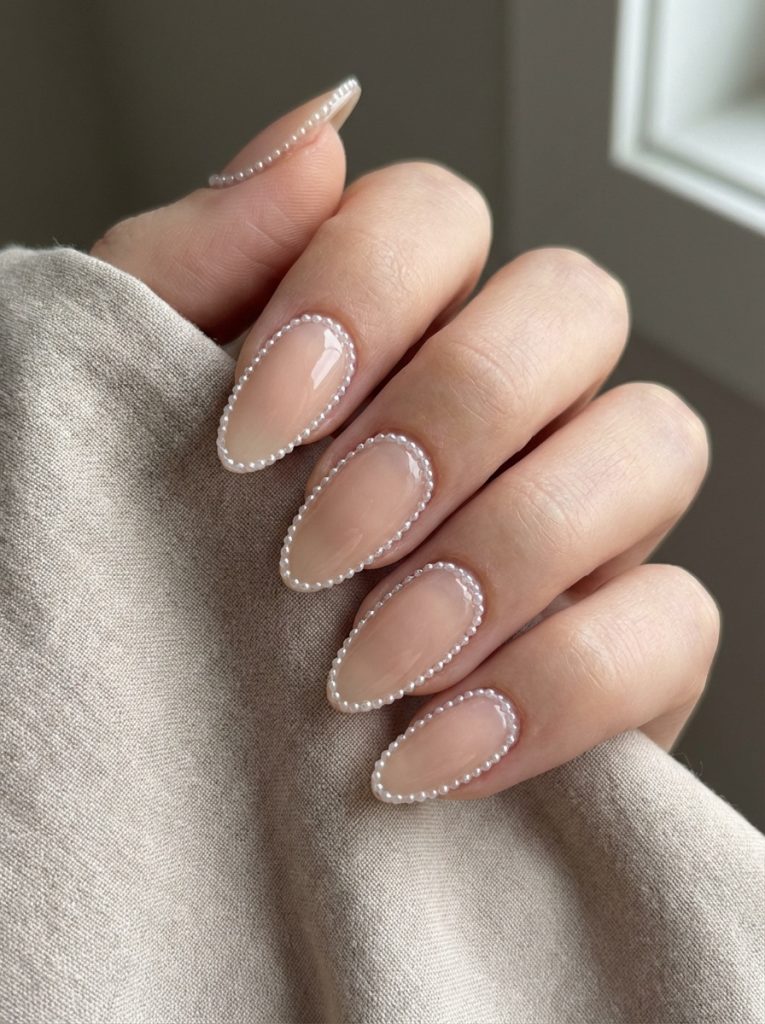 Close-up of almond-shaped nude wedding nails painted creamy beige and bordered with fine pearl beads, photographed against beige fabric near natural light.