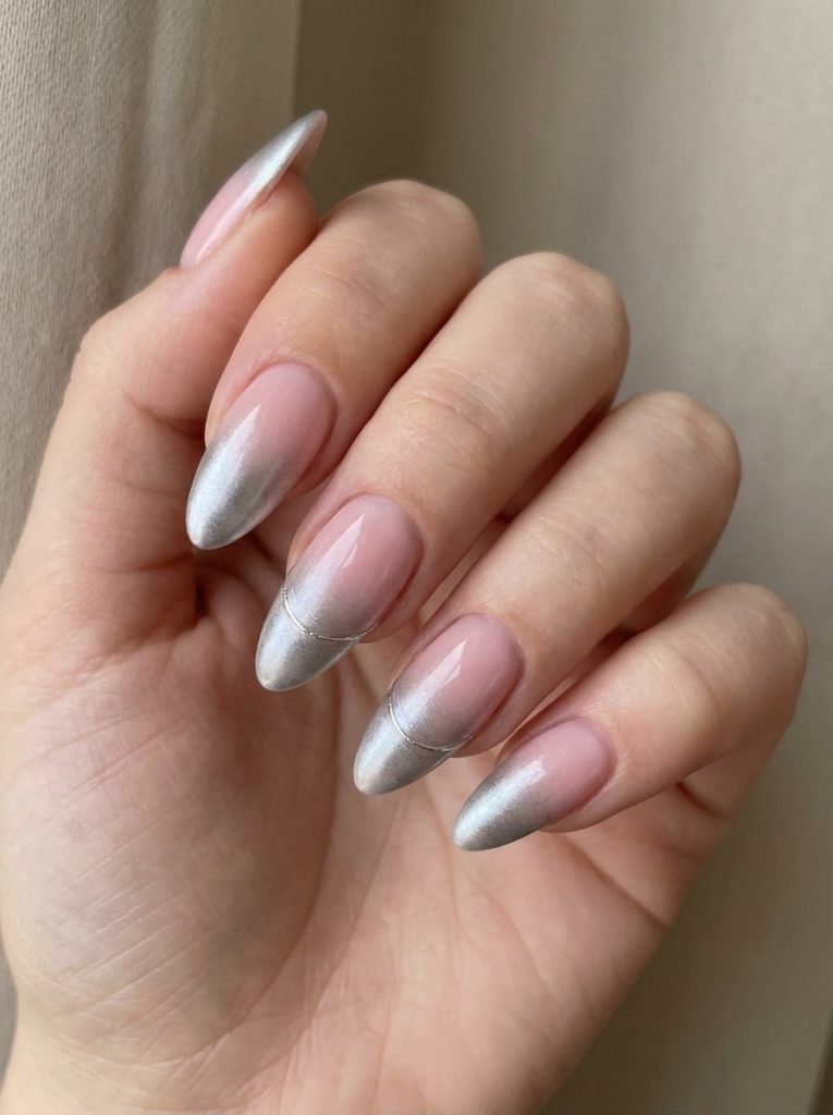 Close-up of a feminine hand near a neutral curtain, showcasing almond ombre wedding nails fading from soft pink into reflective silver chrome tips with a fine curved accent line.