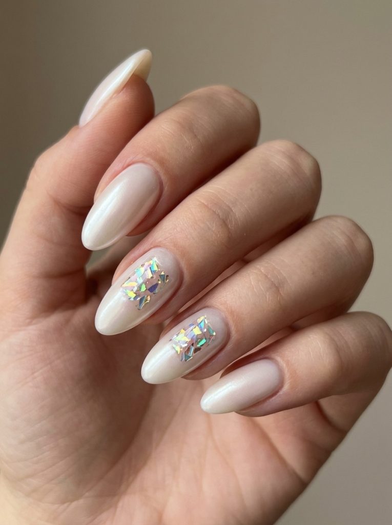 Manicured hand angled inward wearing pearl wedding nails in milky white with holographic shard accents on two elongated almond-shaped nails.