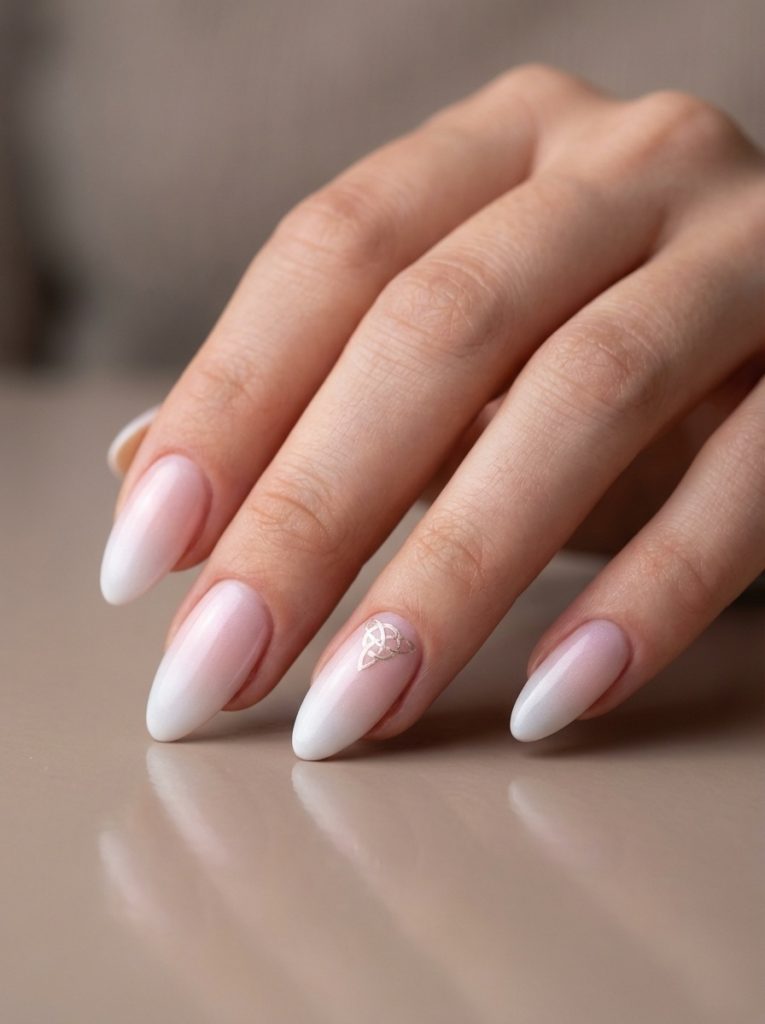 Close-up of a hand resting on a glossy surface featuring almond ombre wedding nails in blush fading to white, with a small metallic knot accent on one nail.