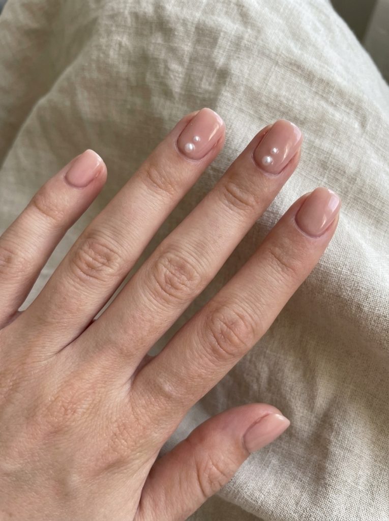 Elegant hand positioned over light linen displaying nude wedding nails in glossy rose-beige polish accented with small round pearls.