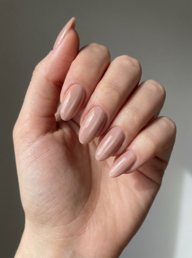 Close-up of a hand showing nude wedding nails in caramel-beige polish with a glossy finish on long almond-shaped nails against a neutral background.