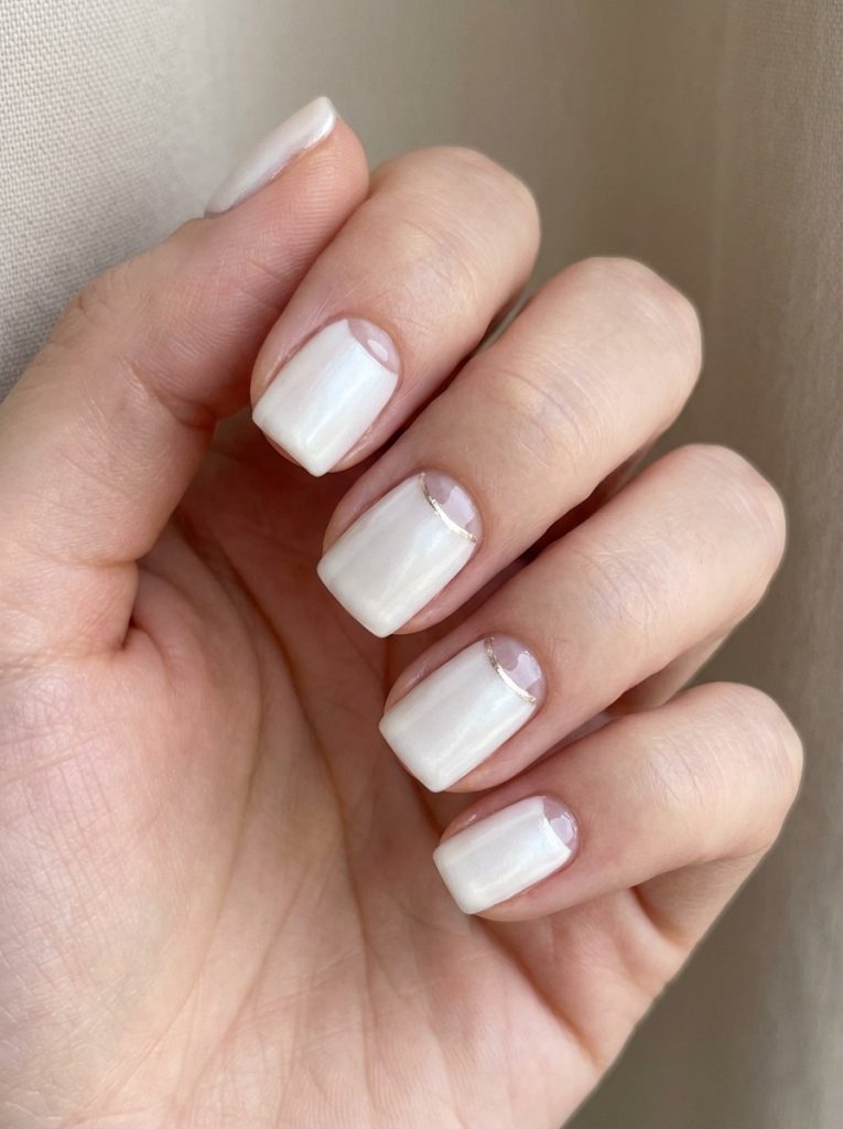 Manicured hand positioned near beige drapery wearing pearl wedding nails in creamy alabaster polish accented by thin gold half-moon detailing at the cuticles.