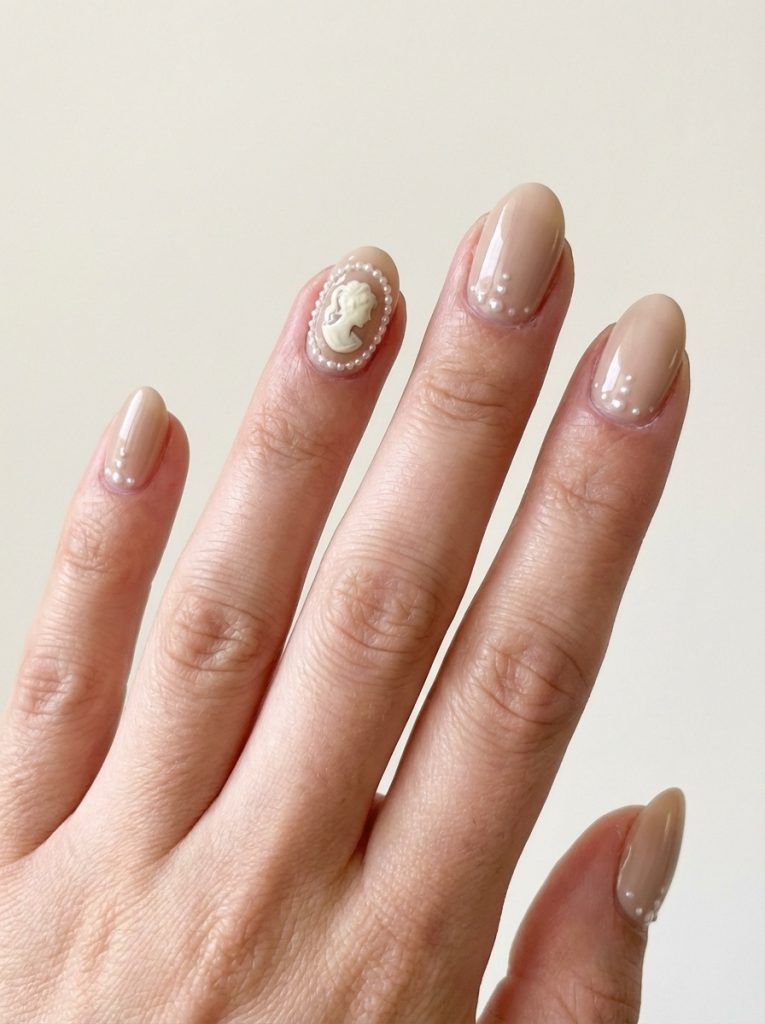 Hand displayed against a soft neutral wall featuring nude wedding nails in warm beige polish with a raised ivory cameo accent surrounded by tiny pearls and dotted cuticle detailing.