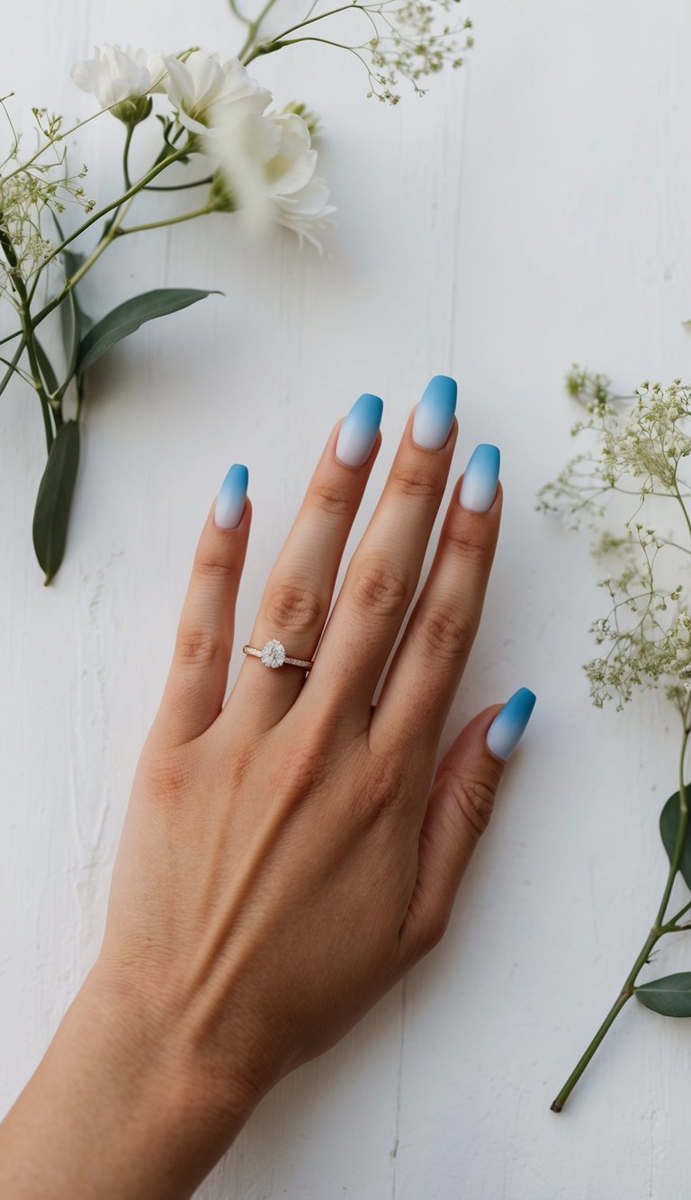 Hand resting on a white wooden surface surrounded by delicate white flowers, featuring ombre wedding nails fading from white into sky blue on long square tips, paired with a diamond ring.