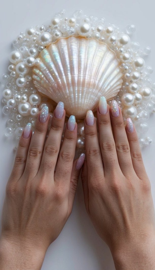 Two hands with almond-shaped pearl wedding nails in pastel ombré shades, featuring subtle shimmer and glitter accents, posed over a large iridescent seashell surrounded by pearls.