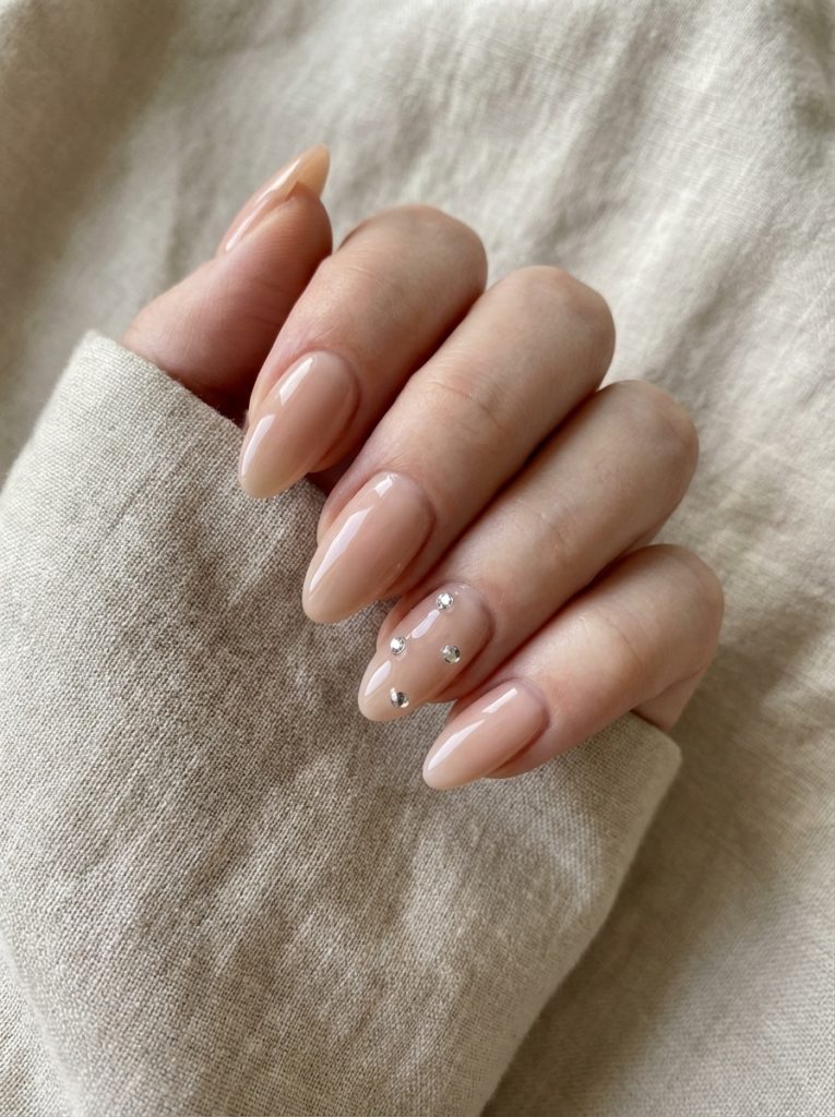 Close-up of a hand resting on linen fabric showcasing nude wedding nails in glossy beige, featuring one accent nail decorated with small silver crystal studs.