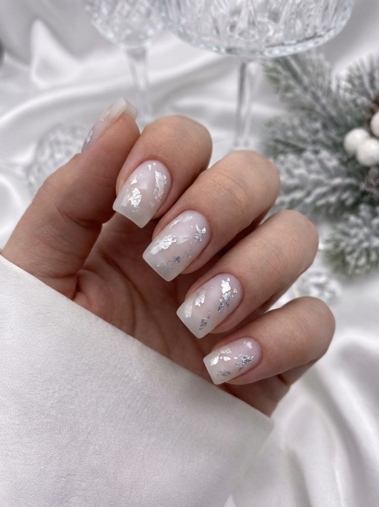 Hand partially wrapped in a smooth satin sleeve displaying milky rose matte wedding nails accented with delicate silver leaf pieces near frosted greenery.