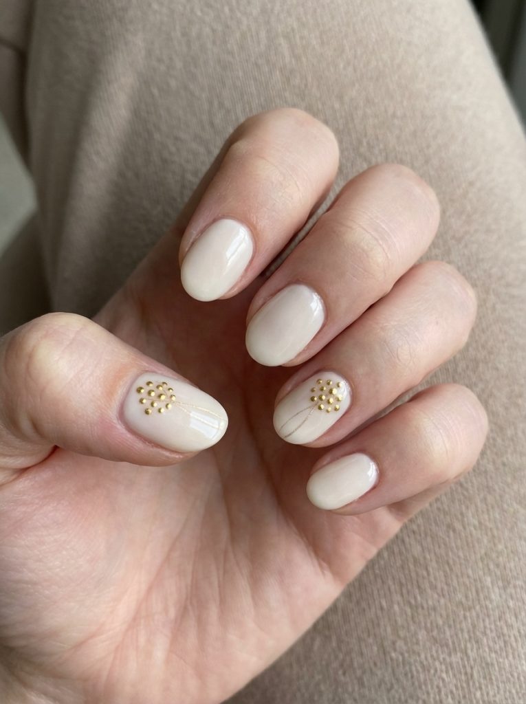 Close-up of short pearl wedding nails painted in warm ivory beige, with tiny gold studs forming delicate floral patterns and fine gold lines on accent nails.